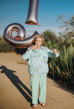 A woman stands on a dirt path with silver balloons, enjoying a joyful moment outdoors in the sun.