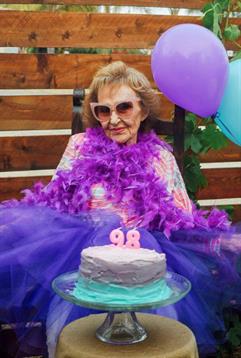 An elderly woman joyfully celebrates her 98th birthday with purple balloons and cake.