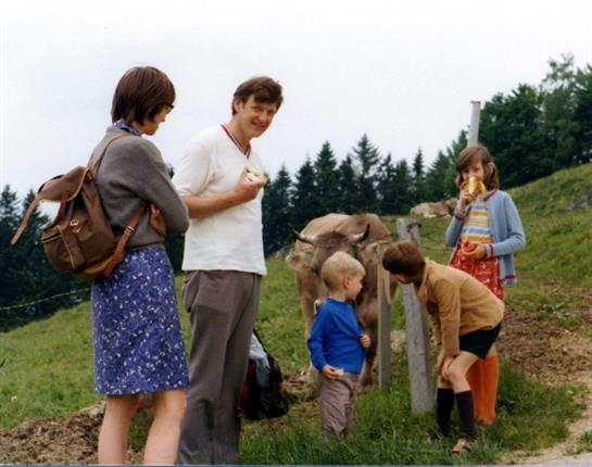 Group of people gather in a green field, engaging with a cow while children play nearby.