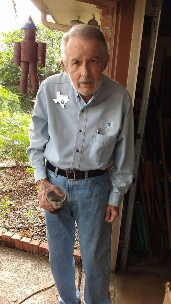 An elderly man wearing a button-up shirt and jeans stands outside, holding a drink.