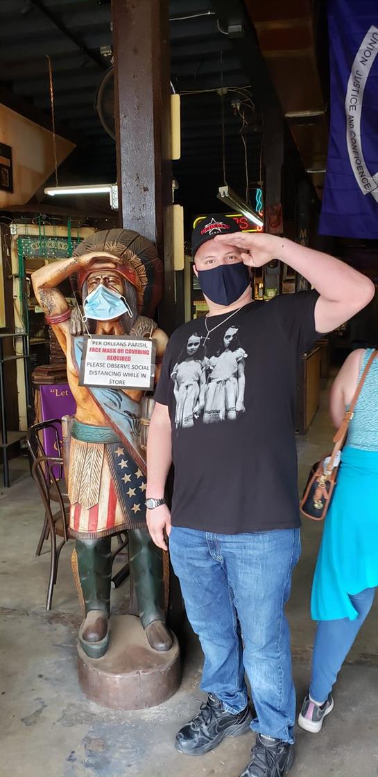 A young man in casual attire salutes a statue wearing a mask, showcasing cultural appreciation.