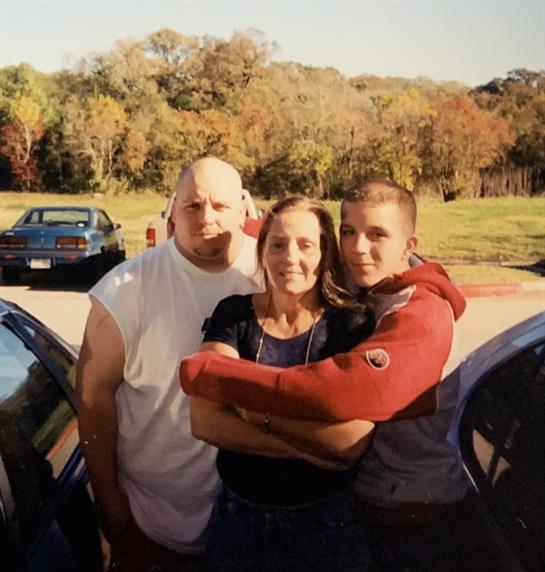 Three friends stand together and smile in a parking lot surrounded by colorful trees.