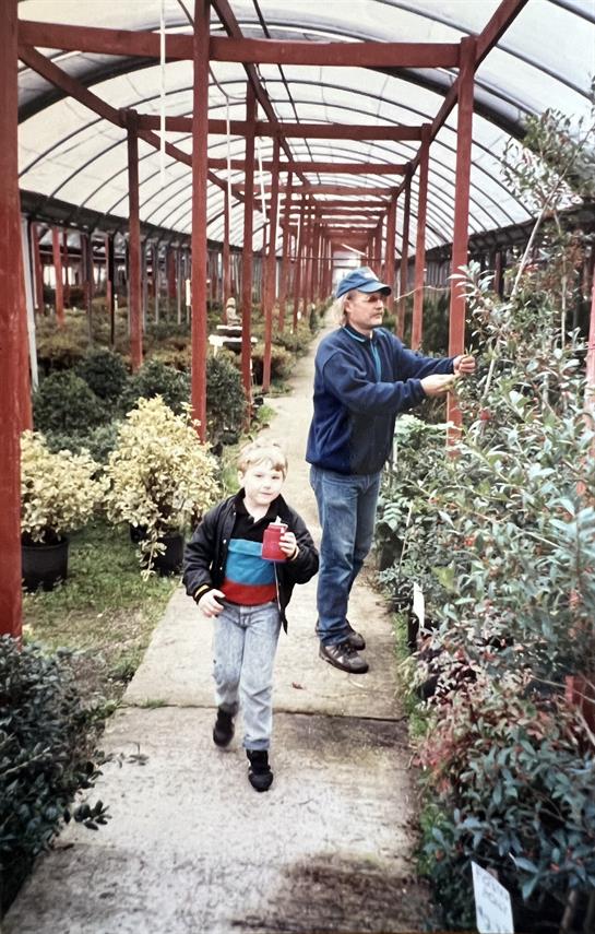 A child walks alongside an adult tending to plants in a greenhouse filled with greenery.
