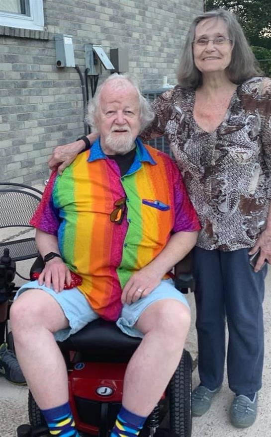 A senior man in a colorful rainbow shirt relaxes outdoors in a wheelchair next to a woman.