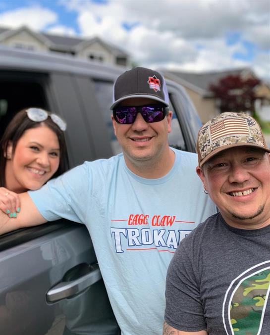 Three friends gather by a parked vehicle, sharing smiles and enjoying a cheerful moment together.