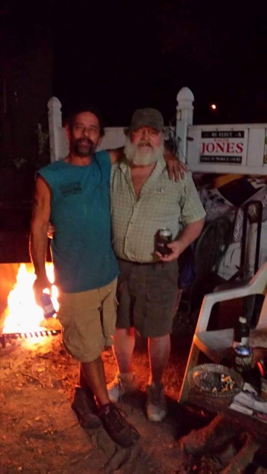 Two friends stand together smiling as they relax by a fire pit, enjoying drinks under the stars.