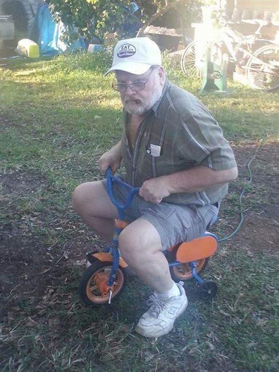 A man with a beard rides a child's tricycle, smiling while sitting outdoors in a green space.