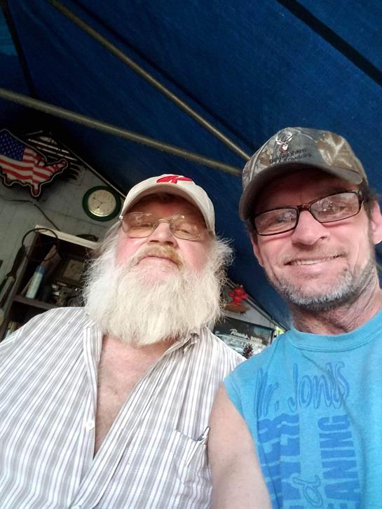 Two men with facial hair and glasses take a cheerful selfie inside a casual bar setting.