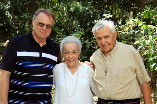 Three family members smile warmly while posing together in a lush, sunny park.