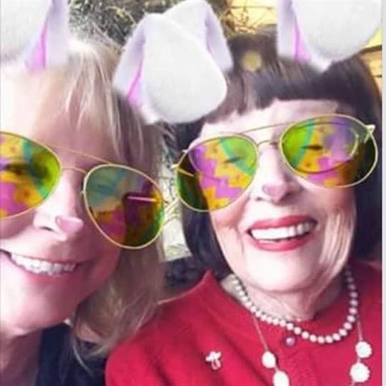 Two women smile brightly while wearing colorful filters at a cheerful event in a lively setting.