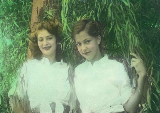 Two young women stand close together, smiling in a vibrant garden filled with green foliage.