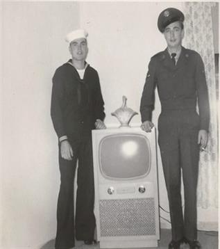 Two servicemen pose next to a vintage television in a simple indoor space, showcasing their attire.