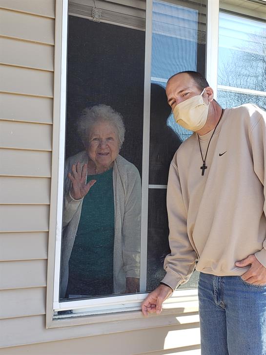 A young man stands outside a house, smiling while the elderly woman waves at him from inside.