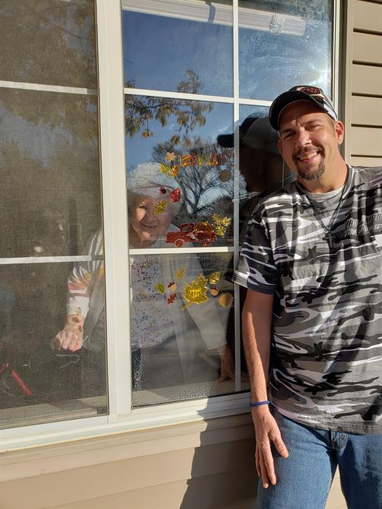 A man stands next to a window, smiling, while a woman is playfully peeking from behind the glass.