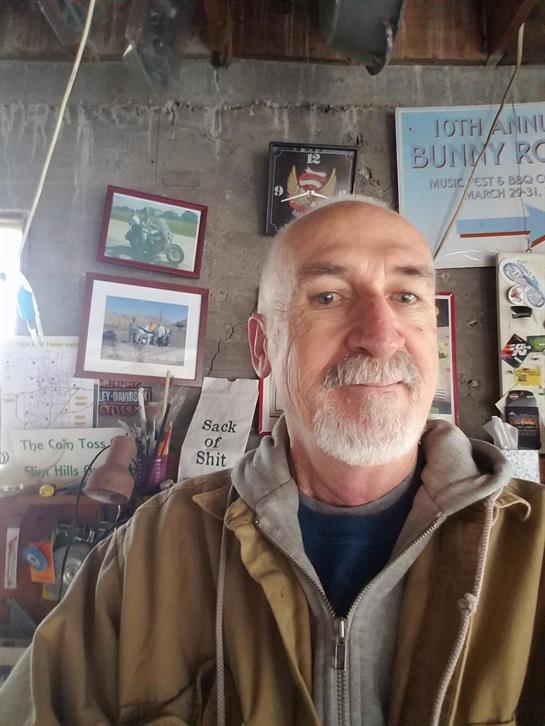 A man smiles while posing in a rustic workshop filled with vintage signs and tools.