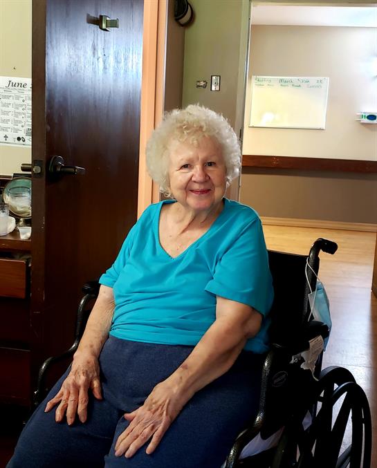 A cheerful elderly woman in a turquoise shirt is seated in her wheelchair inside a care facility.