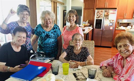 Five elderly women smile and share stories while enjoying refreshments in a bright kitchen.