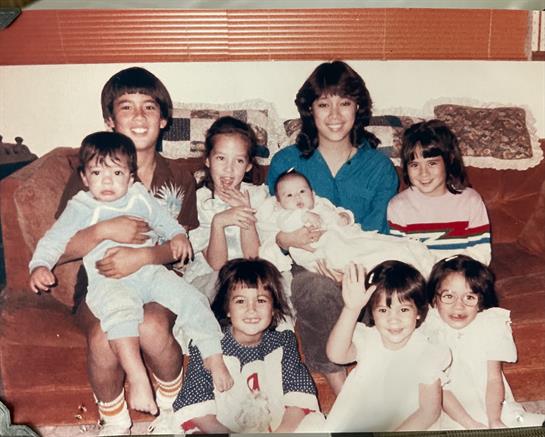 A group of eight children and teenagers smiles and interacts on a couch in a living room.