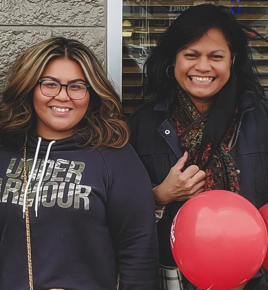 Two joyful friends pose with red balloons outside a local store, showcasing their happy expressions.
