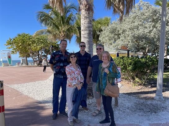 Five friends gather together in a beachside park surrounded by palm trees and sunlight.
