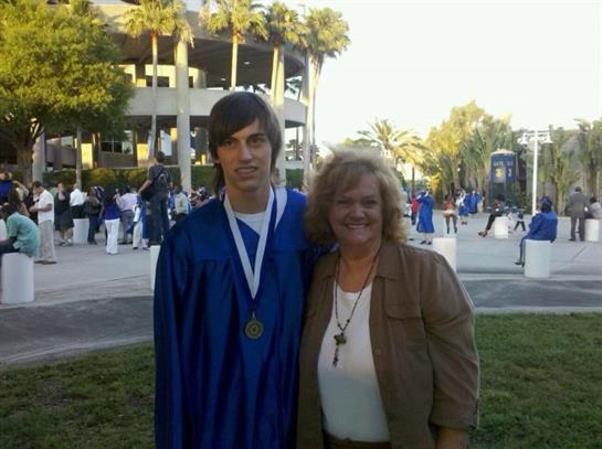 A young graduate in blue robe stands with a family member, both smiling in a sunny outdoor setting.
