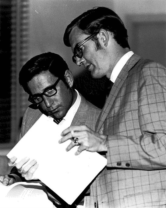 Two suited men review papers together, engaged in conversation about important matters.