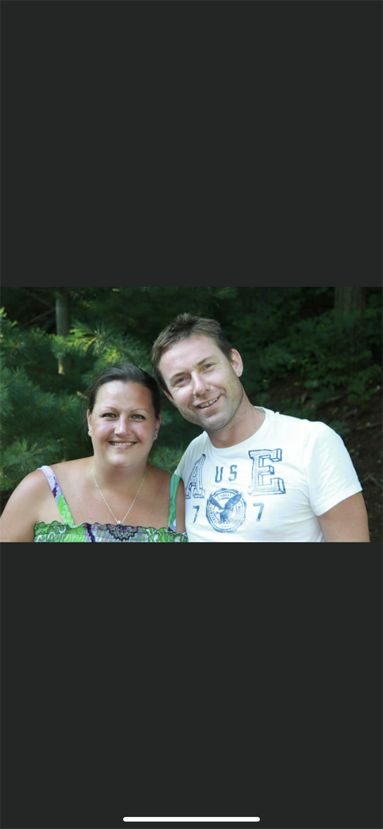 Smiling couple stands together in a vibrant, green landscape on a sunny day.