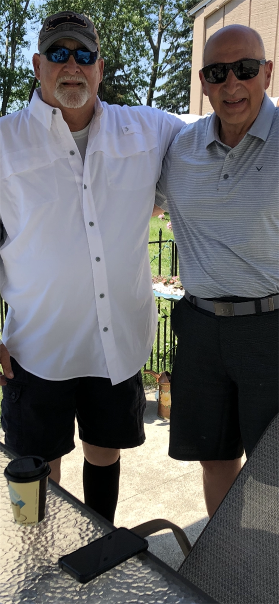 Two men stand close together smiling while wearing casual clothes in a bright outdoor setting.