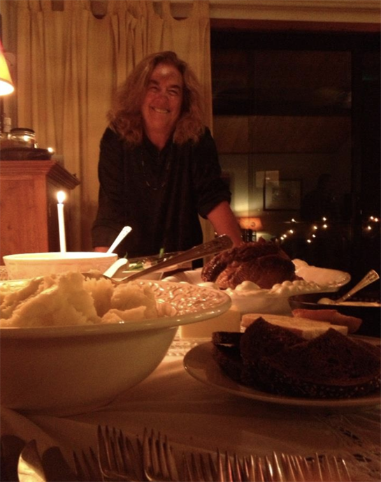 A person stands happily behind a table filled with various delicious dishes in soft candlelight.