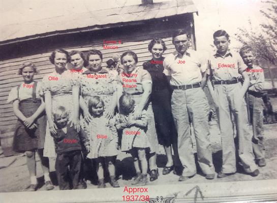 Several family members stand together in front of a rustic wooden house, showcasing togetherness.