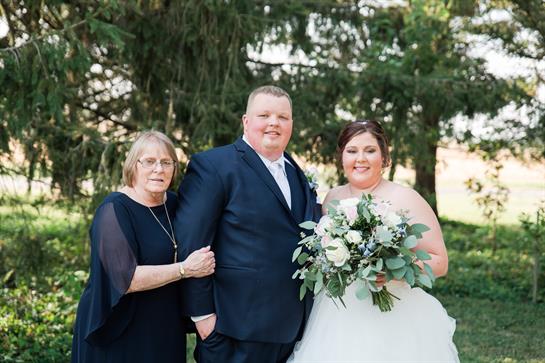 A bride holds a bouquet while standing with her parents in a lush garden backdrop.