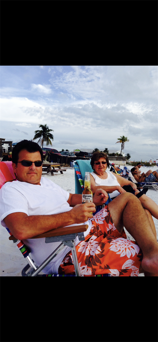 Two adults enjoy drinks while lounging on beach chairs by the shore, surrounded by palm trees.