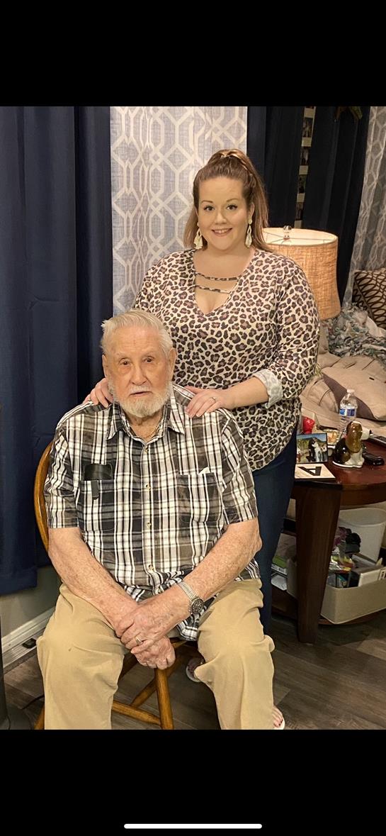 Elderly man in plaid shirt sits while woman in spotted top stands behind him, both smiling.