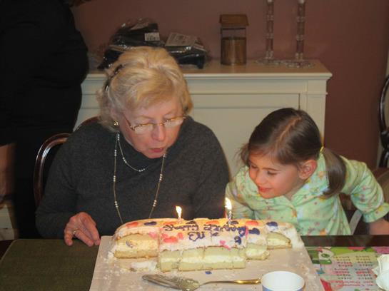 A grandmother and granddaughter blow out candles on a birthday cake in a cozy indoor setting.