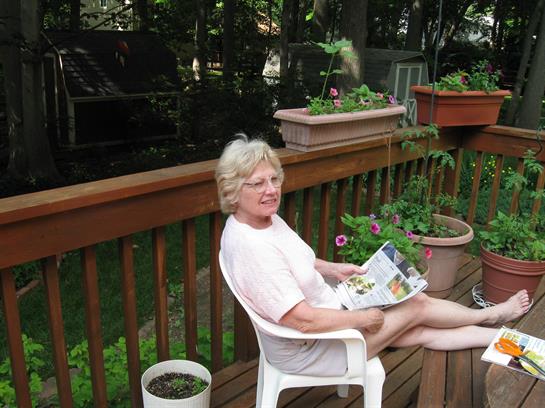A senior woman sits comfortably on a deck chair, enjoying a magazine amid vibrant flower pots.
