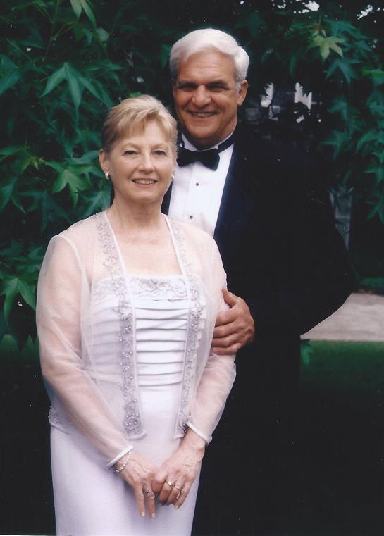 A couple poses happily together in formal attire, surrounded by vibrant greenery.