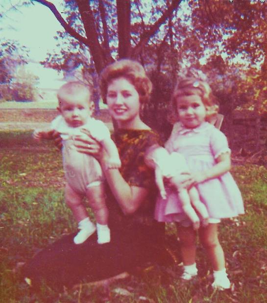 A mother holds her baby while her daughter stands beside them, both smiling happily in a garden.