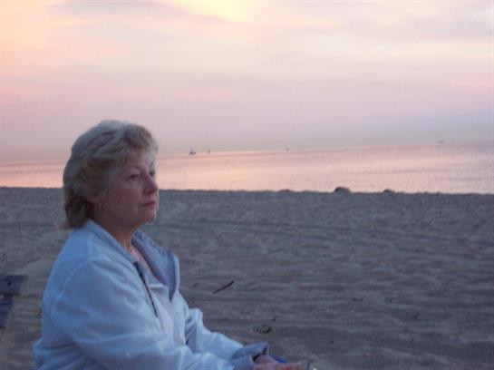An older woman sits quietly on the beach, reflecting as the sun sets over the horizon.