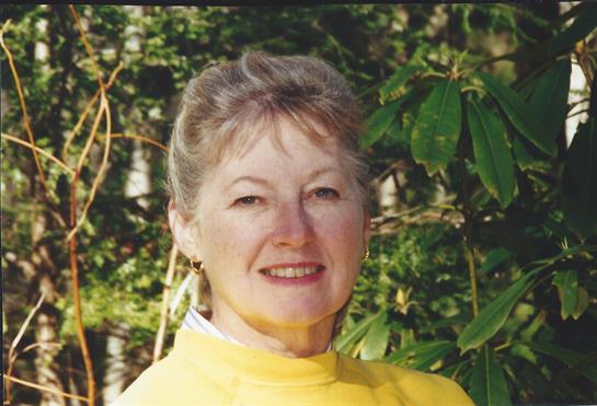 A woman with a warm smile stands outdoors amid vibrant green foliage in the sun.