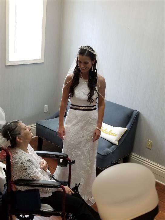 A bride in a white dress smiles at her grandmother while getting ready indoors.