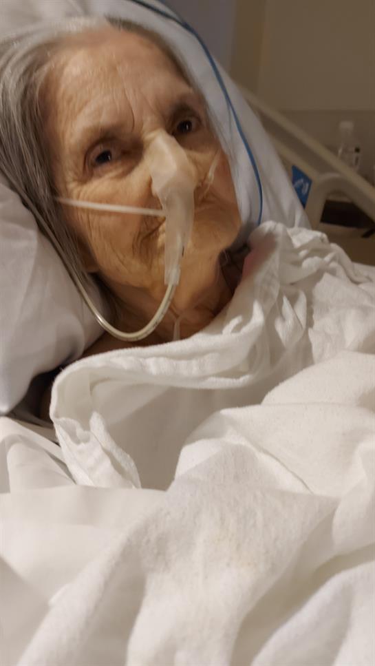An elderly woman, connected to medical equipment, rests in a hospital bed while receiving care.