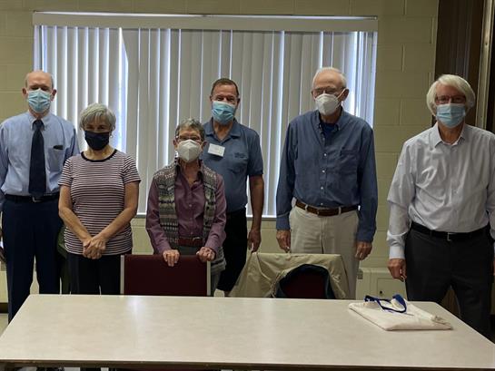 Several people wearing masks stand together in a community center, engaging in a meeting activity.