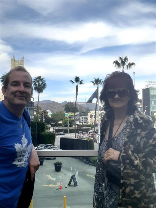 Two people stand on a balcony overlooking a vibrant landscape with palm trees and mountains.