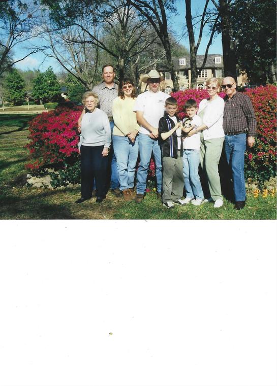 A group of family members poses happily in front of vibrant floral bushes on a clear day.