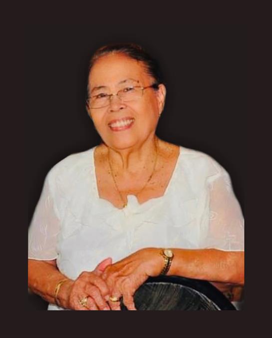 An elderly woman in glasses smiles warmly while seated at a gathering.