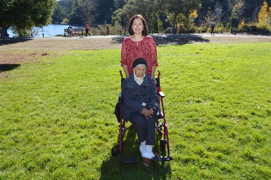 A woman standing behind another in a wheelchair smiles in a sunny park setting by a lake.