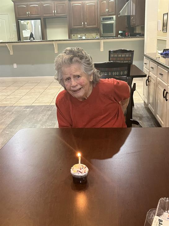 An elderly woman smiles at a table with a cupcake and candle, celebrating a special day.