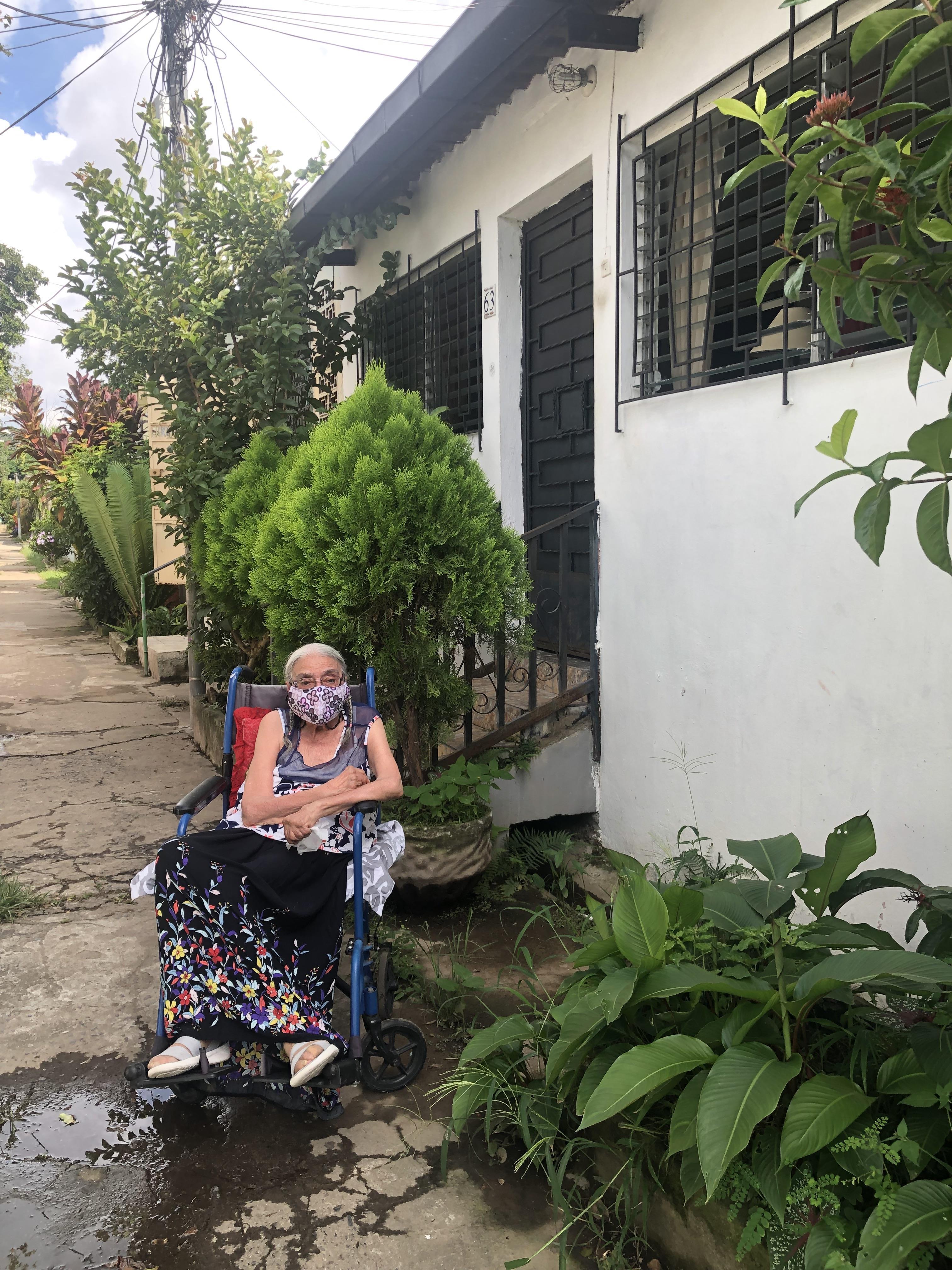 An elderly woman relaxes in a wheelchair outside her house on a sunny day, wearing a mask.