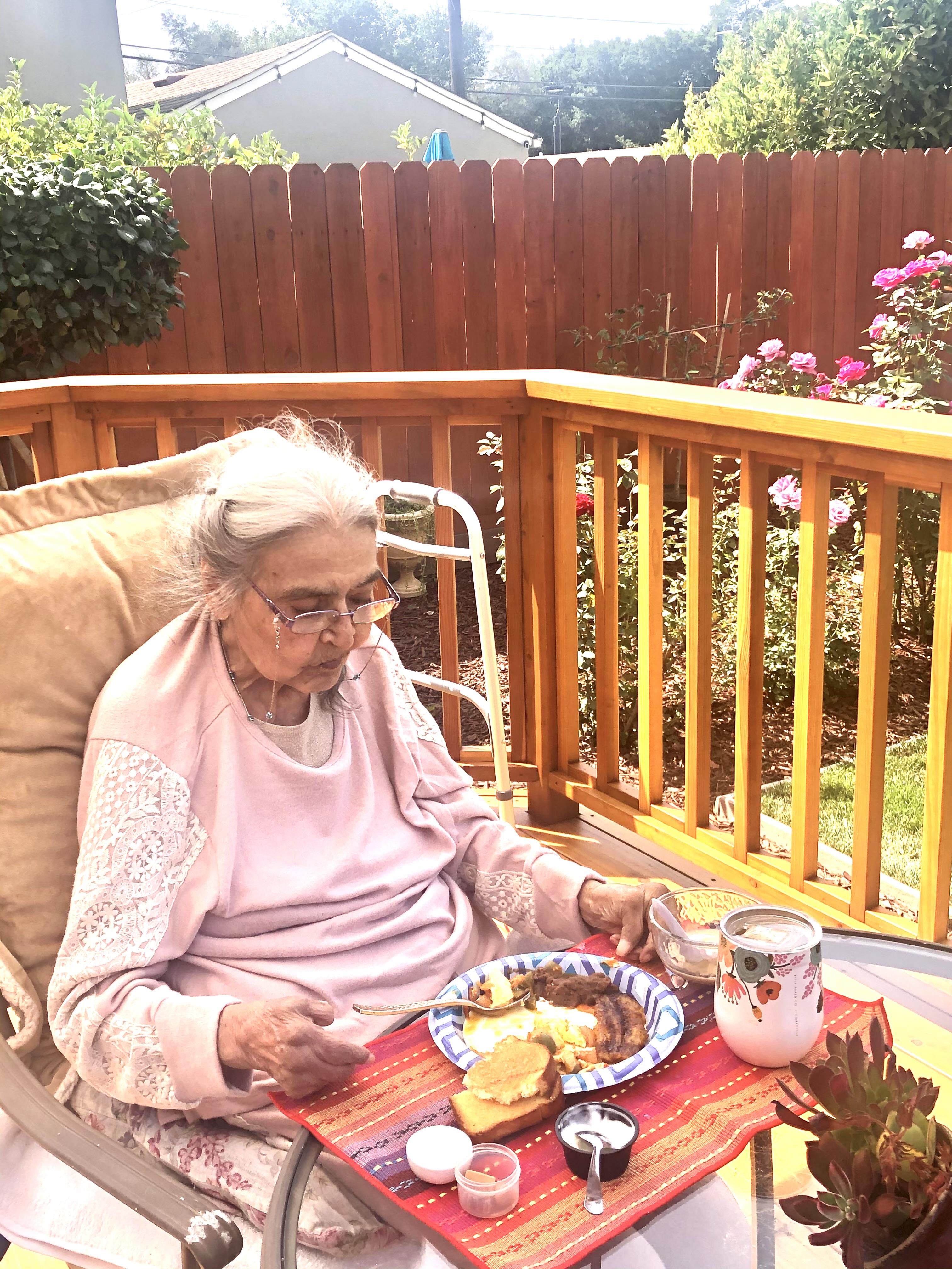 Elderly woman sits on a patio chair, savoring breakfast with a peaceful garden view.
