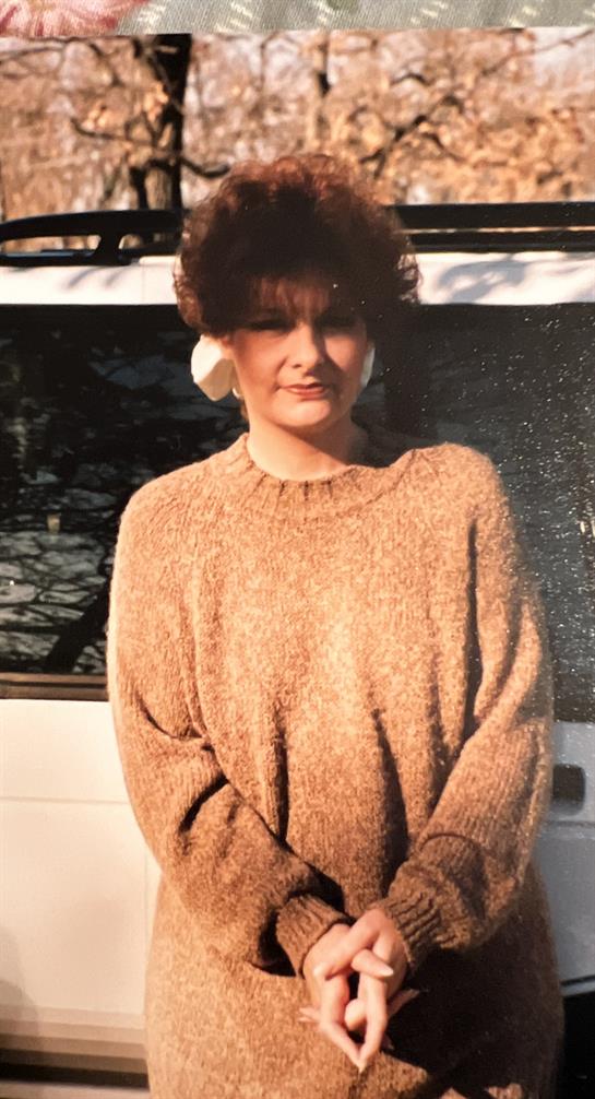 A woman stands smiling, wearing a warm sweater, near a parked vehicle under the sunlight.
